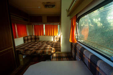 Esta imagen muestra el interior de una furgoneta camper de estilo retro con tapicería a cuadros, cortinas naranjas, una cama acogedora, armarios superiores y la luz del sol entrando por la ventana.