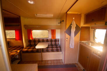 Esta imagen muestra el acogedor interior de una autocaravana, con asientos a cuadros, una mesita, una cocina compacta, cortinas naranjas y la luz del sol entrando por las ventanas.