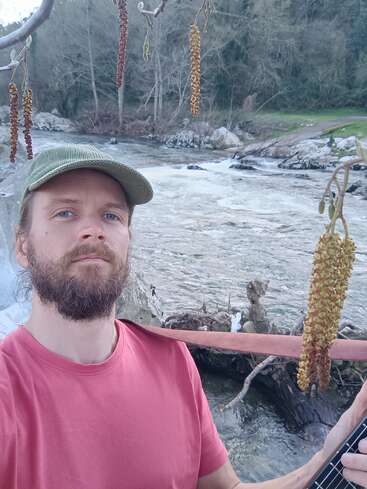 Un hombre barbudo, con una camisa rosa y una gorra verde, está de pie junto a un río, con una guitarra en la mano. Los árboles y los amentos colgantes enmarcan el paisaje natural al aire libre.