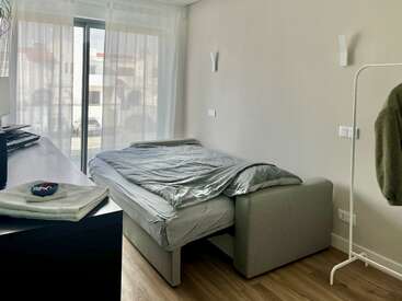 Un dormitorio luminoso y minimalista con un sofá cama gris, cortinas transparentes, una cómoda, un teclado electrónico, un perchero, una toalla y suelo de madera, que transmite un ambiente acogedor y moderno.