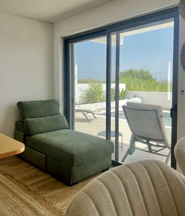 Un espacio acogedor y moderno cuenta con una chaise longue verde situada junto a unas puertas correderas de cristal que dan a un patio soleado con tumbonas, una pequeña piscina y una exuberante vegetación en el exterior.