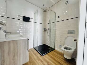 Este cuarto de baño cuenta con una mampara de ducha de cristal, azulejos blancos modernos con una raya negra, suelo de madera, mueble de lavabo blanco, inodoro suspendido y toalla negra. Diseño minimalista.