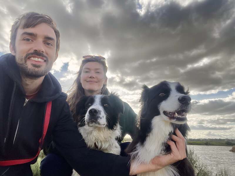La imagen muestra a un hombre y una mujer posando con dos perros blancos y negros en un entorno al aire libre, con el hombre a la izquierda y la mujer en el centro.