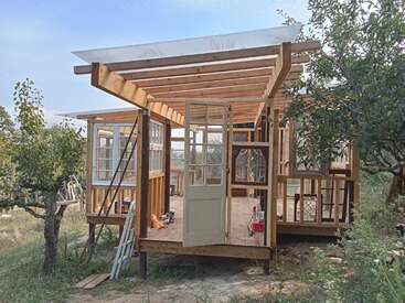 Esta imagen muestra una casa de madera en construcción, con vigas vistas, armazón abierto, múltiples ventanas y árboles que la rodean en un entorno natural al aire libre. Se ven herramientas.