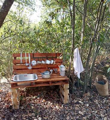 La imagen muestra una cocina al aire libre, con un fregadero, ollas y utensilios sobre una mesa de madera, rodeada de árboles y hojas, y una toalla colgada cerca.