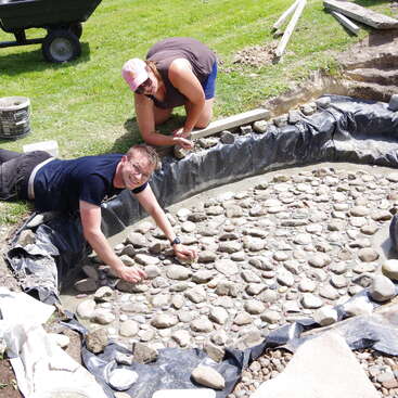 Ein Mann und eine Frau bauen an einem sonnigen Tag einen Teich mit Steinen, umgeben von Gras und Baumaterialien.