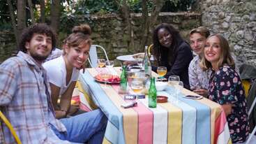 Five friends enjoy an outdoor meal together around a colorful table, smiling and laughing, with drinks, snacks, and a rustic stone wall in the background.