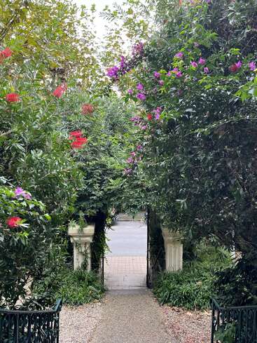 A garden path framed by lush greenery and vibrant pink flowers leads to a small gate, opening onto a quiet street beyond, surrounded by nature’s beauty.