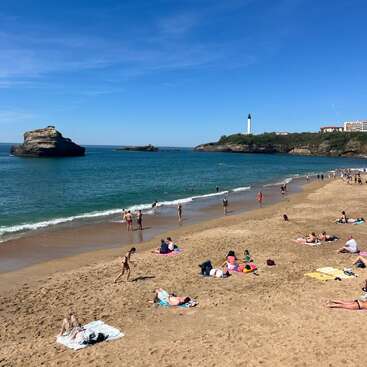 People relax on a sandy beach under a clear blue sky. Some swim in the sea, while a lighthouse stands in the distance on a rocky outcrop.