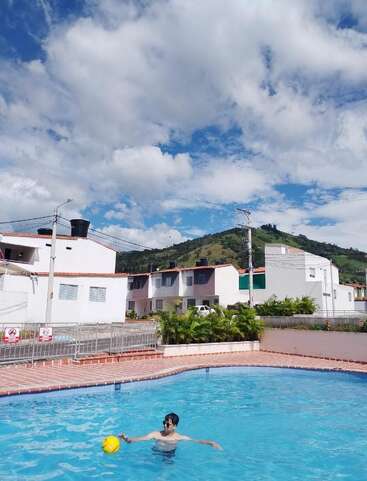 Jemand hat Spaß im Pool mit einem gelben Ball. Im Hintergrund sorgen weiße Häuser, grüne Hügel, blauer Himmel und flauschige Wolken für eine lebendige, entspannte Outdoor-Szene.
