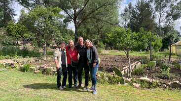 Vier Menschen stehen lächelnd in einem üppigen Garten zusammen, umgeben von grünen Bäumen, Pflanzen und einem Gemüsebeet. Die Atmosphäre ist sonnig, friedlich und entspannt.