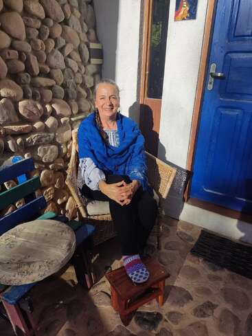 The image depicts a woman sitting on a wicker chair outside a house, with a blue door and stone wall in the background, bathed in sunlight.