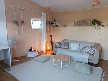 Esta sala de estar aconchegante apresenta um sofá bege com almofadas estampadas, plantas penduradas, uma luminária de chão brilhante, um tapete, uma mesa de centro, uma escrivaninha e arte decorativa na parede.