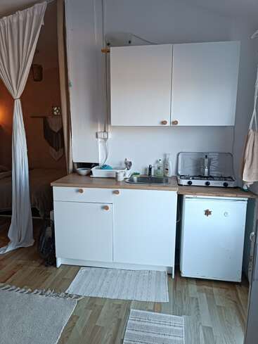 Esta imagem mostra uma área de cozinha pequena e aconchegante com armários brancos, uma pia, um fogão, um frigobar, piso de madeira clara e decoração simples e minimalista.