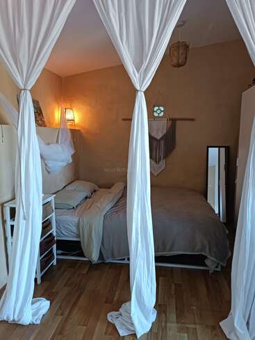 Um quarto aconchegante apresenta uma cama com roupa de cama clara, iluminação suave, cortinas brancas transparentes, uma pequena mesa de cabeceira, piso de madeira, decoração de parede e uma lâmpada decorativa.