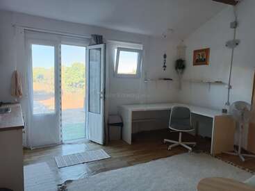 Uma sala iluminada com piso de madeira, uma grande janela e uma porta aberta. Há uma mesa branca em forma de L, uma cadeira de escritório, uma pequena área de cozinha e decorações mínimas.