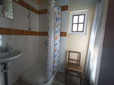 Esta imagem mostra um banheiro pequeno com uma área de banho, uma cortina de chuveiro colorida, uma pia, uma cadeira de madeira, uma pequena janela e azulejos brancos.