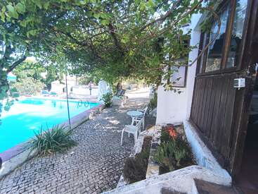 Um pátio externo sereno com paralelepípedos, sombreado por videiras verdes exuberantes, tem vista para uma piscina transparente. Cadeiras brancas e charme rústico criam uma atmosfera relaxante.