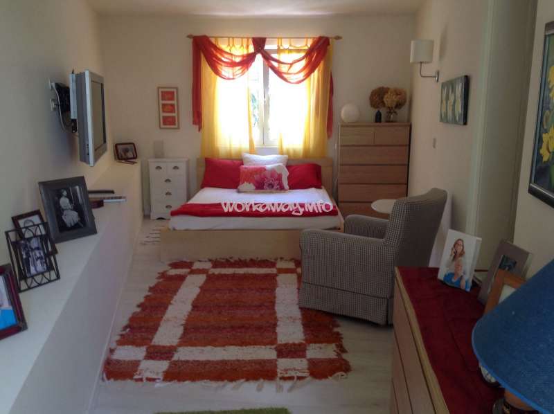 Cette chambre confortable est agrémentée d'un lit aux touches rouges, de rideaux jaunes, d'un tapis orange, d'un fauteuil, d'une commode, d'une télévision, de photos encadrées et d'une décoration chaleureuse et accueillante.