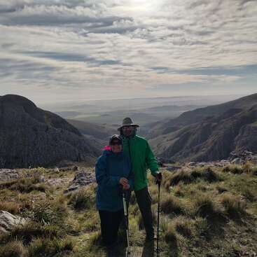 Dois caminhantes estão juntos em uma encosta de montanha gramada, cercada por penhascos rochosos impressionantes, sob um céu nublado e ensolarado, desfrutando de uma paisagem cênica.