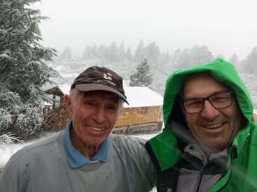 Zwei lächelnde Männer machen ein Selfie im Freien in einer verschneiten Winterlandschaft. Einer trägt eine Brille und eine grüne Kapuze, der andere eine dunkle Baseballmütze. Gemütlicher Moment.