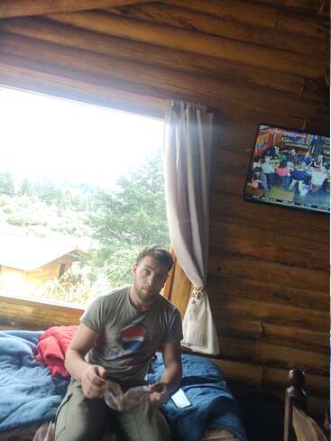 Ein Mann sitzt auf einem Bett in einer gemütlichen Blockhütte und hält einen Gegenstand in der Hand. Ein großes Fenster zeigt die Bäume draußen, während ein Fernseher an der Holzwand steht.