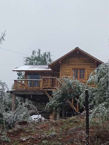 Eine gemütliche Holzhütte steht erhöht zwischen schneebedeckten Bäumen und hat einen Balkon mit Blick auf eine winterliche Landschaft. Leichter Schneefall schafft eine ruhige, friedliche und rustikale Atmosphäre.