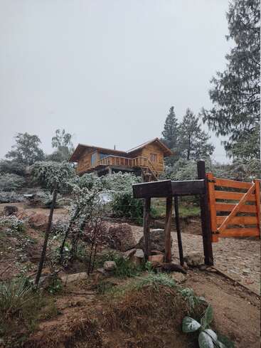 Eine charmante Holzhütte liegt auf einem kleinen Hügel, umgeben von schneebedeckten Bäumen und Sträuchern, und ein rustikales Tor markiert den Eingang.