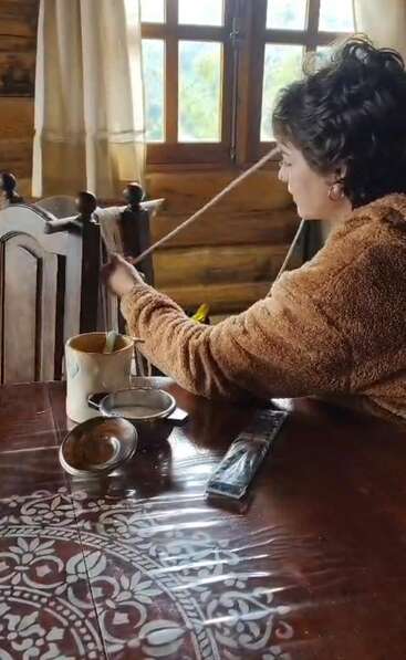 Eine Frau sitzt an einem Holztisch und spinnt mit der Hand Garn. Das Sonnenlicht fällt durch das Fenster. Tassen, Teller und Utensilien stehen auf dem gemusterten Tisch.