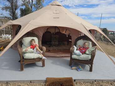 Duas crianças relaxam em cadeiras confortáveis em frente a uma grande tenda bege. O clima parece agradável, e há brinquedos espalhados pelo chão nas proximidades.