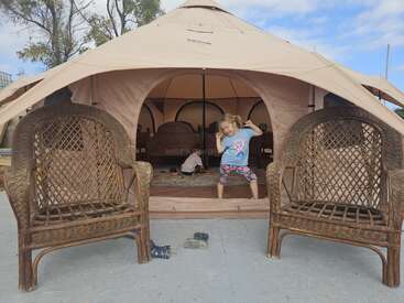 Duas cadeiras de vime estão sentadas em frente a uma grande tenda bege. Dentro, duas crianças brincam no chão, com camas visíveis ao fundo. Céu azul acima de você.
