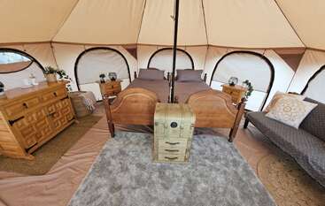 Esta imagem mostra uma aconchegante barraca de glamping com duas camas de madeira, mesinhas de cabeceira, uma cômoda, um sofá, um baú e uma decoração rústica e aconchegante, além de plantas.