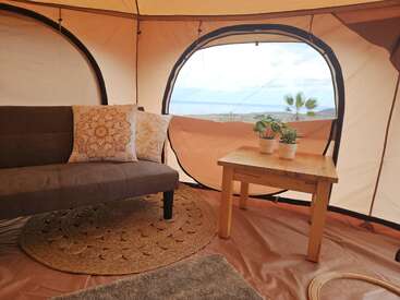 Dentro de uma tenda aconchegante, um sofá marrom com almofadas estampadas fica ao lado de uma mesa de madeira com vasos de plantas, com vista para uma paisagem externa. Atmosfera acolhedora.