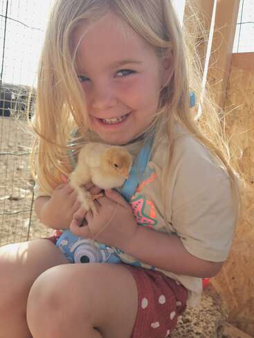 A smiling young girl with long blonde hair gently holds a fluffy yellow chick. She wears a cartoon camera necklace and red polka dot shorts, enjoying sunshine.
