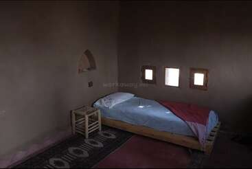 Dieses Bild zeigt ein einfaches, rustikales Zimmer mit einem Einzelbett, einem kleinen Holzhocker, erdfarbenen Wänden, drei kleinen Fenstern und gemusterten Teppichen auf dem Boden.