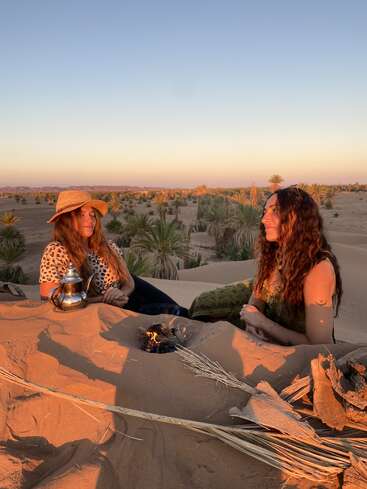 Zwei Frauen sitzen bei Sonnenuntergang auf sandigen Dünen, umgeben von Palmen. Sie unterhalten sich und trinken Tee aus einem Kessel, während zwischen ihnen ein kleines Feuer brennt. Friedliche Atmosphäre.