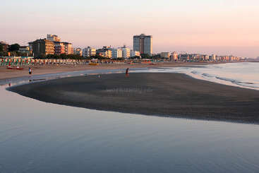 La imagen representa una serena escena de playa con un mar en calma, en la que aparecen una orilla arenosa y unos cuantos individuos, probablemente tomada durante el día, lo que transmite una sensación de tranquilidad y relajación.