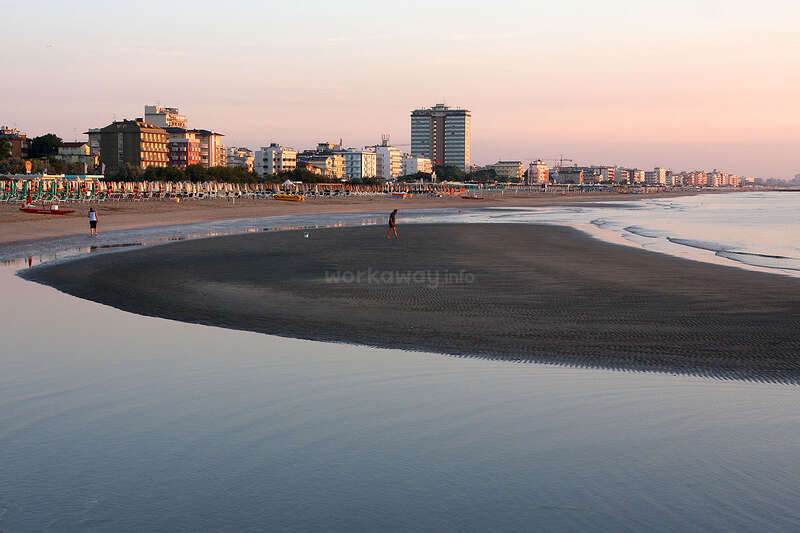 La imagen representa una serena escena de playa con un mar en calma, en la que aparecen una orilla arenosa y unos cuantos individuos, probablemente tomada durante el día, lo que transmite una sensación de tranquilidad y relajación.