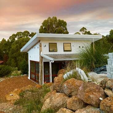 The image depicts a modern white house with a flat roof, situated on a hillside amidst large rocks and trees, set against a cloudy sky.
