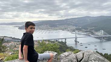 A boy sits on rocks overlooking a scenic river, bridge, and town below. Green hills, houses, and a cloudy sky create a peaceful, breathtaking landscape view.
