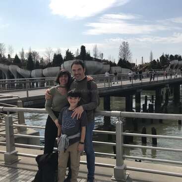 A family of three poses happily by a waterfront railing, with a bridge, water, and a modern park structure in the background under a partly cloudy sky.