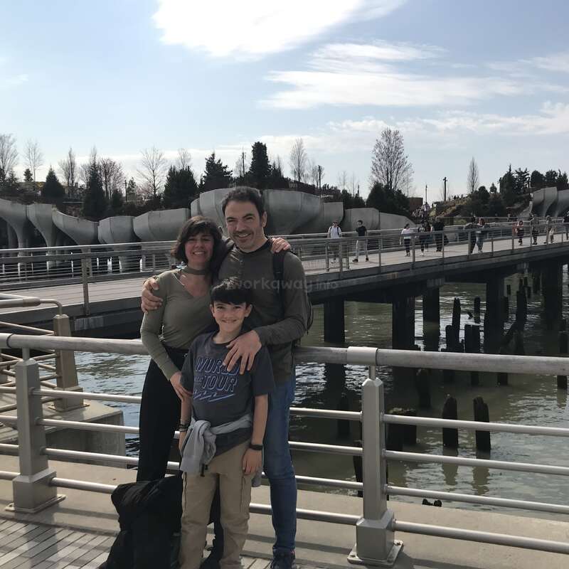 A family of three poses happily by a waterfront railing, with a bridge, water, and a modern park structure in the background under a partly cloudy sky.