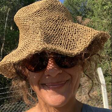 Uma pessoa sorridente usa um grande chapéu de palha trançada e óculos escuros. Ela tem um piercing no nariz e está ao ar livre, cercada por vegetação e luz solar.