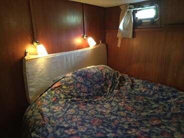 The image depicts a small, dimly lit room with a bed, featuring a floral-patterned comforter, a white headboard, and two wall-mounted lights, set against a wood-paneled backdrop.