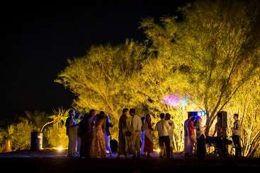 A group of people gathers outdoors at night, illuminated by warm lights among tall trees. The scene feels festive, social, and relaxed, with soft, natural surroundings.