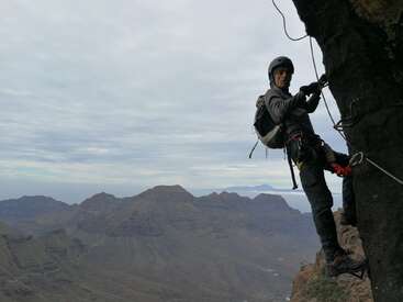 Um homem está escalando uma rocha em um penhasco íngreme, usando capacete, arnês e cordas, com uma paisagem montanhosa ao fundo sob um céu nublado.