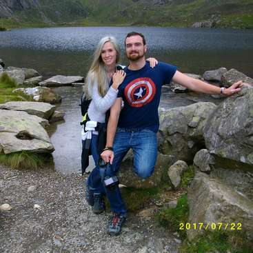 Das Bild zeigt einen Mann und eine Frau, die vor einem See für ein Foto posieren. Der Mann trägt ein Captain America-T-Shirt und die Frau hat lange blonde Haare.