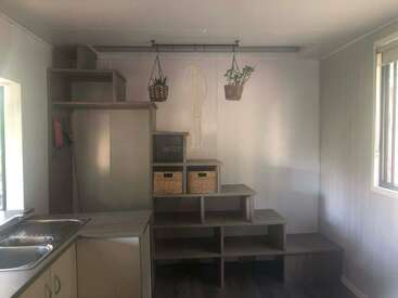 This image shows a small, minimalist room with a kitchenette on the left, a staircase with storage boxes, hanging plants, a macrame, and ample natural light.