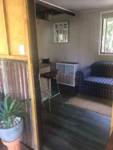 A cozy room with a blue plaid couch, small desk, shelving unit, and a plant by the door. Light streams in through a window.