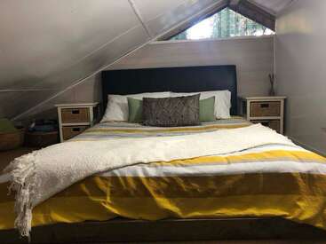 This cozy attic bedroom features a comfortable bed with yellow-striped bedding, throw pillows, two nightstands with wicker drawers, a window, and a white angled ceiling.